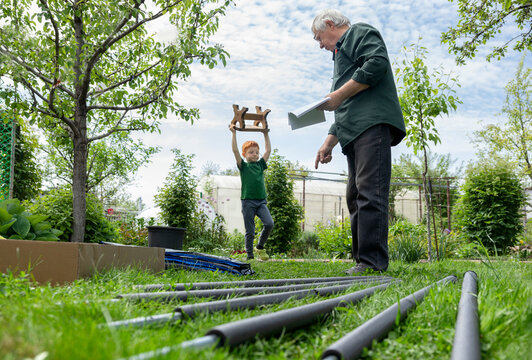 An elderly man reads instructions while a boy carries a stool to help assemble a trampoline outdoors