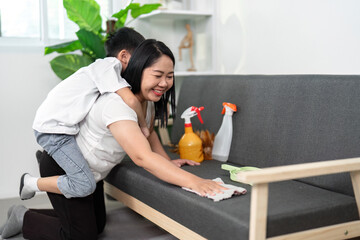 A parent cleans a couch while a child playfully rides on their back, showcasing joy in household...