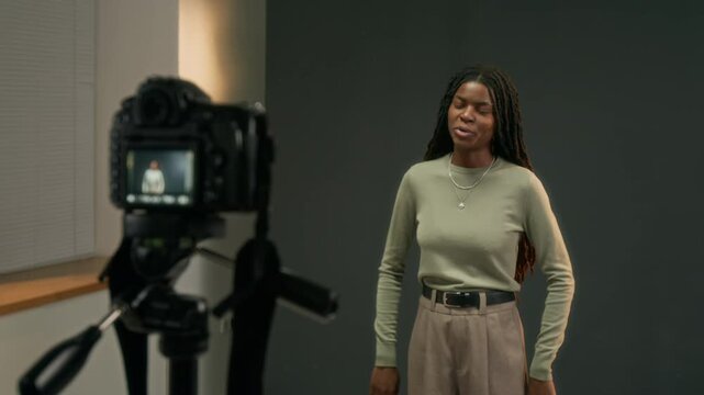 African American woman standing against gray backdrop and acting on tripod camera in studio