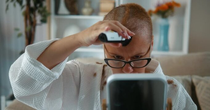 Determined and confident adult woman with cancer, after chemotherapy, is cutting her hair, shaving her head with a hair clipper at home in the living room.