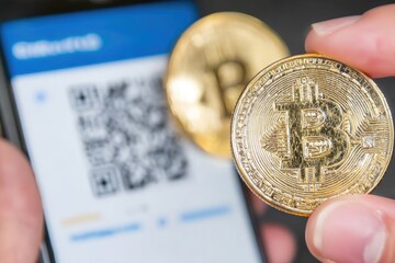 Hand holding a physical Bitcoin coin next to a QR code on a phone