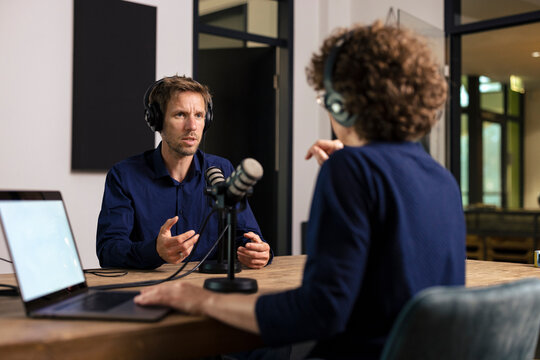 Man wearing headset discussing with presenter sitting at desk in recording studio