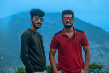 Two young men with beards and glasses pose confidently  against a misty mountain backdrop. The...