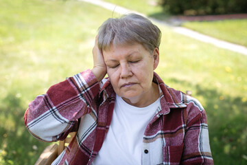 Elderly woman in red plaid shirt holding head in pain while sitting in sunny garden, concept of...