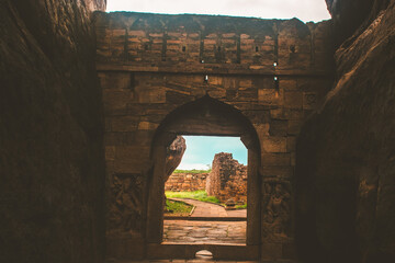 A historic arched gateway carved from stone, opening toward a bright green valley beyond —...