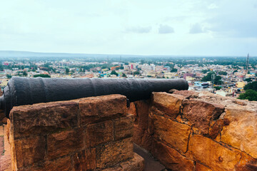 An old cannon rests on the ramparts of a historical Indian fort, aimed toward the sprawling cityscape below.