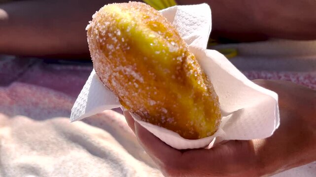 Hand holding a malasada donut with sugar at a beach food stand, close-up of fast food in colorful wrapper, yellow bikini