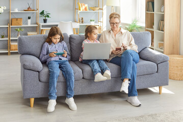 Smiling grandmother sitting on sofa with two grandchildren using digital devices. Older woman...