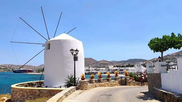 Video of a scenic coastal view of a traditional white windmill on the island of Paros, Greece, overlooking the vibrant blue Aegean Sea and the charming port of Parikia.