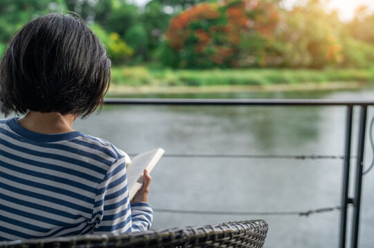Woman sitting on chair reading a book on balcony overlooking river during golden hour morning. Self care, digital detox, and mindful slow living. Emotional wellness in riverside retreat. Quiet luxury.