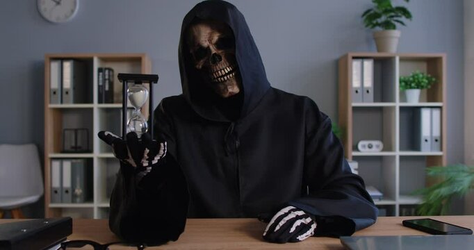 Demonic devilish nightmare fantasy Grim Reaper in spooky skull mask dark hooded cloak disguise at office work desk with hour glass reminds to fear inevitably approaching deadline. Death, time metaphor