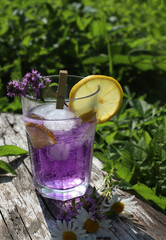 On the street on a wooden stump in transparent glasses a purple cocktail is poured with a slice of lemon and a sprig of thyme