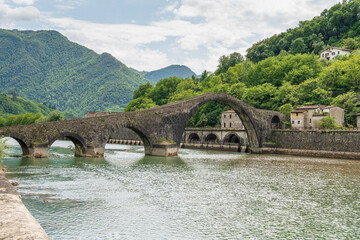 Stone Devil's Bridge over river with green hills and old buildings in Tuscany, Italy.