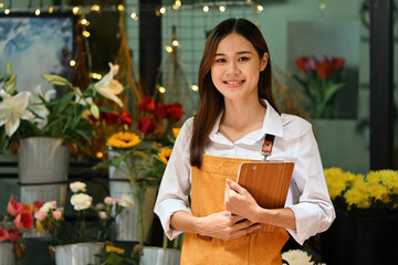 A cheerful young entrepreneur standing confidently in her flower shop, holding a clipboard and...