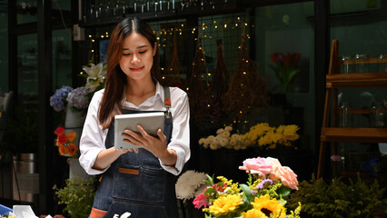 An Asian female florist using a digital tablet to manage flower shop operations, surrounded by colorful blooms