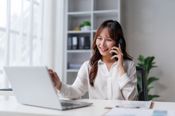 Young Asian businesswoman engaging in a phone conversation while working on a laptop in a bright, modern office setting