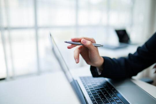 Businesswoman holding a pen and pointing at a laptop screen while engaged in work at a bright, modern office environment