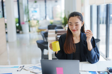 Young Asian businesswoman drinking coffee and working on laptop in modern office, analyzing...