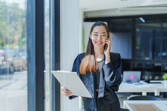Smiling Asian businesswoman talking on the phone while holding a clipboard, working efficiently in a modern office environment - Powered by Adobe