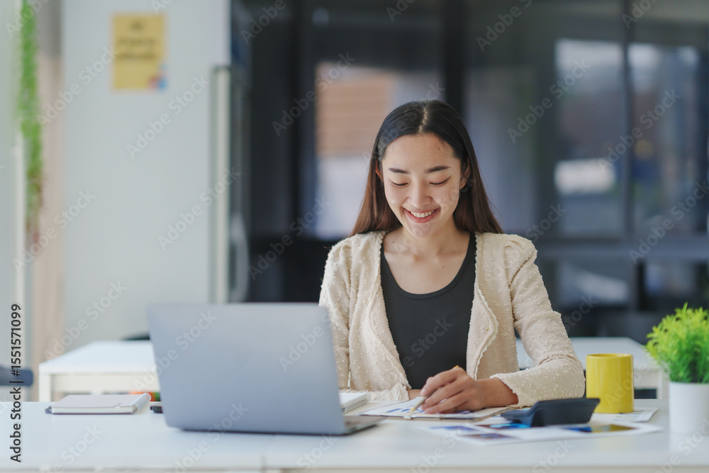 Poster smiling asian businesswoman analyzing marketing data and taking notes while working on laptop comput