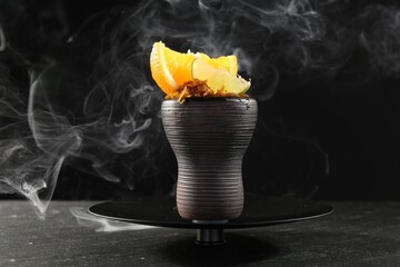 Hookah bowl with citrus fruits and smoke on black table, closeup