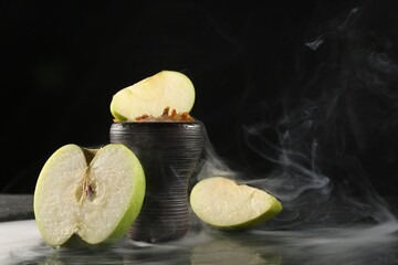 Hookah bowl with apple pieces and smoke on black table, closeup. Space for text