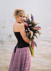 A girl in a beautiful dress walks on the shore of a lake with a bouquet of lupines