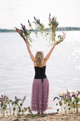A girl in a beautiful dress walks on the shore of a lake with a bouquet of lupines