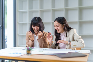 Two asian businesswomen are celebrating successful project looking at financial report, raising...