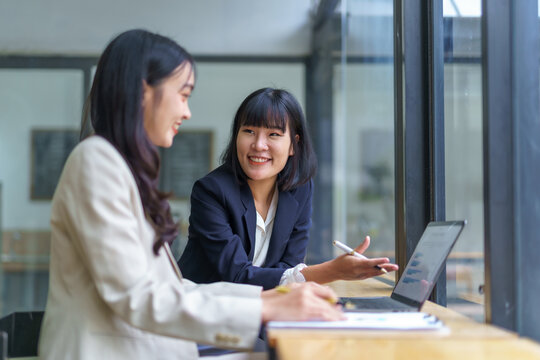 Two asian businesswomen discussing marketing strategy using laptop and taking notes in modern office