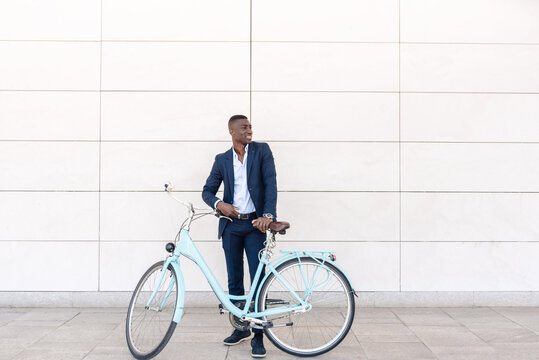 African american businessman standing with bicycle in front of a white tiled wall and looking away