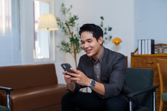Asian businessman sitting comfortably in a modern office waiting area, using a smartphone for browsing and staying connected