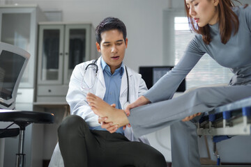 Asian doctor examining a female patient's injured ankle while she rests in a hospital bed, focusing...