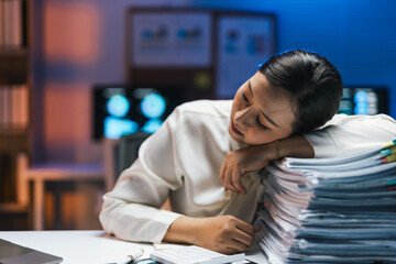 Asian office worker resting head on a pile of paperwork, tired after working overtime late at night in office