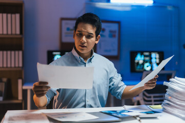 Young professional working late in a modern office, comparing and analyzing documents on a computer screen, managing a heavy workload