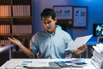 Young businessman looking confused and stressed while reviewing paperwork and working late at night in office