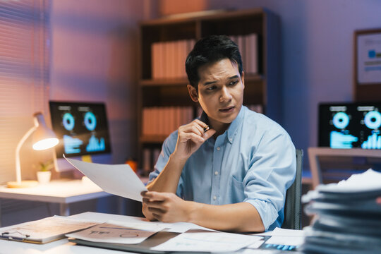 Focused asian financial analyst reading documents and analyzing statistics displayed on computer screen working late at night in office