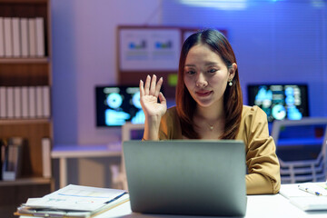 Young Asian businesswoman waving during a video call on her laptop in a modern office, illuminated by evening lights