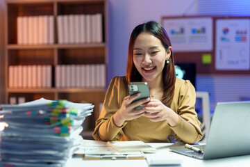 Happy businesswoman using mobile phone while working late at night in office with documents and laptop on desk