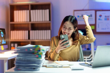 Asian female entrepreneur feeling happy and raising her arm while reading good news on her smartphone in office at night