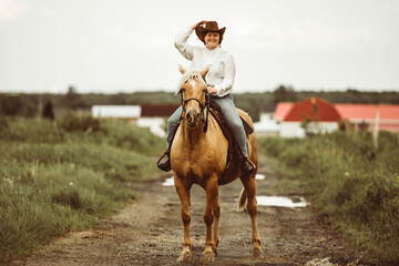 A woman wearing a cowboy hat is riding a horse along a dusty dirt road