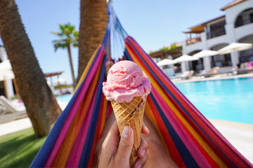 POV: Relaxing in a colorful hammock with a pink ice cream cone by a resort pool. Embodying the perfect summer vacation, leisure, and tropical indulgence.