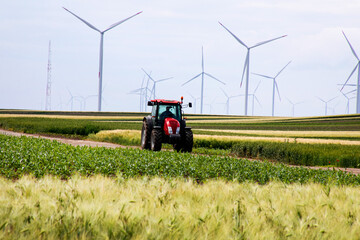 Red tractor traversing verdant fields beneath towering wind turbines, celebrating Green Energy Day...