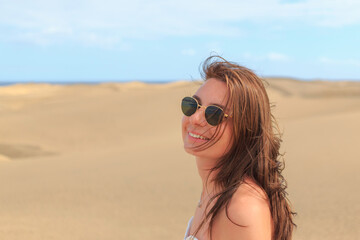 A joyful woman stands in the sandy dunes, wearing sunglasses and a breezy outfit, with wind-swept hair and a big smile against the blue sky.