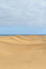 Expansive sand dunes stretch towards the horizon, merging with a tranquil ocean under a partly...