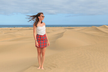 A young woman in a plaid skirt and top enjoys the warm breeze while standing barefoot in a sandy desert, with ocean waves visible in the background.