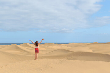 A person stands barefoot on golden sand dunes, arms raised in celebration, with the ocean visible in the background under a partly cloudy sky.