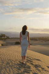A woman walks barefoot on sandy dunes as the sun sets, casting warm colors across the landscape and highlighting the peaceful atmosphere.