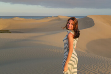 A woman in a striped dress walks gracefully on the sandy dunes as the sun sets, creating a serene ambiance in the coastal landscape.