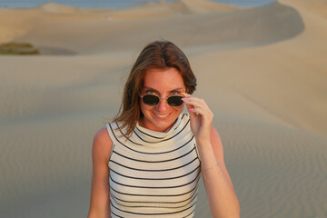A woman with sunglasses smiles while standing on golden sand dunes during a warm afternoon in a remote desert landscape.